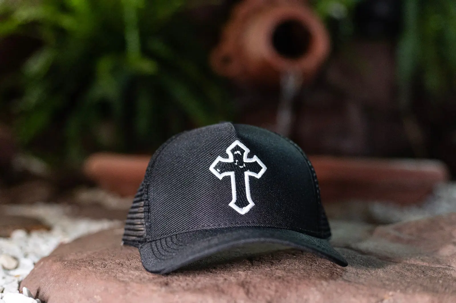 Boné da Cruz preto, com a cruz bordada em branco, sobre uma pedra
