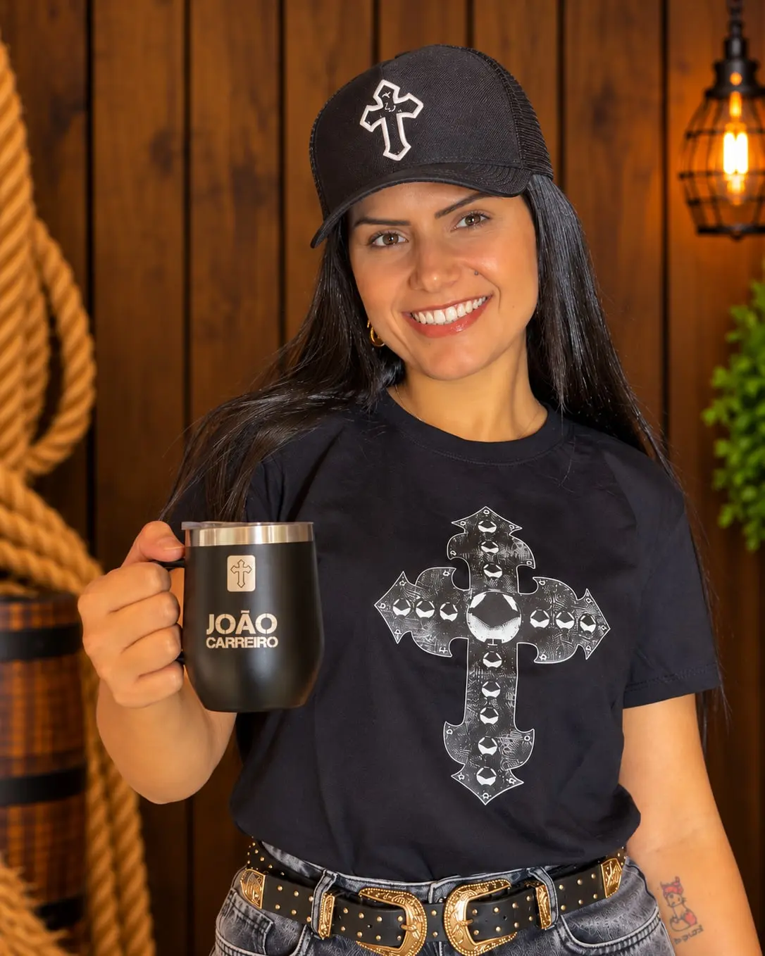 Modelo sorrindo, de boné preto, com a Camiseta Cruz do Chapéu e a caneca João Carreiro na mão